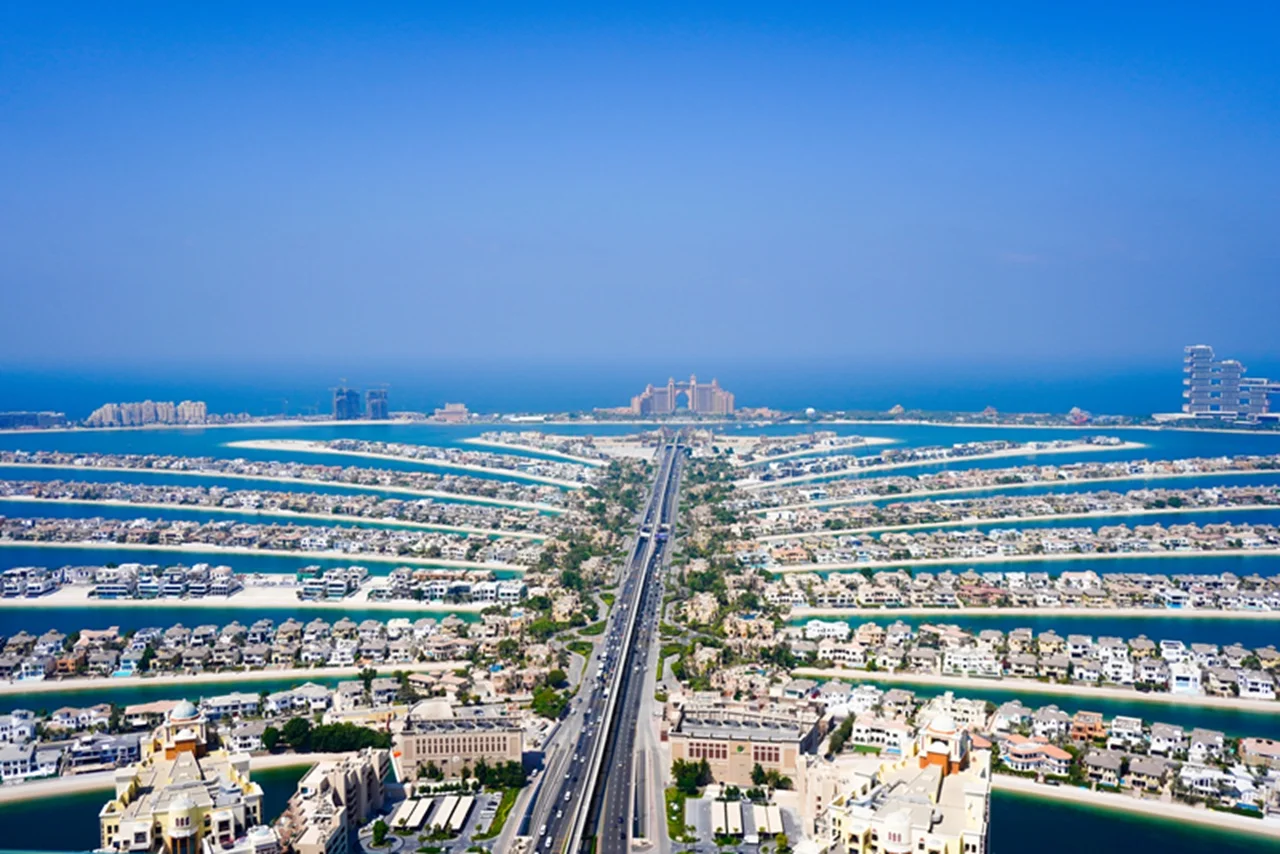 Aerial view of Palm Jumeirah, Dubai, featuring a central highway flanked by palm-shaped residential areas. Surrounding blue sea creates a serene atmosphere.