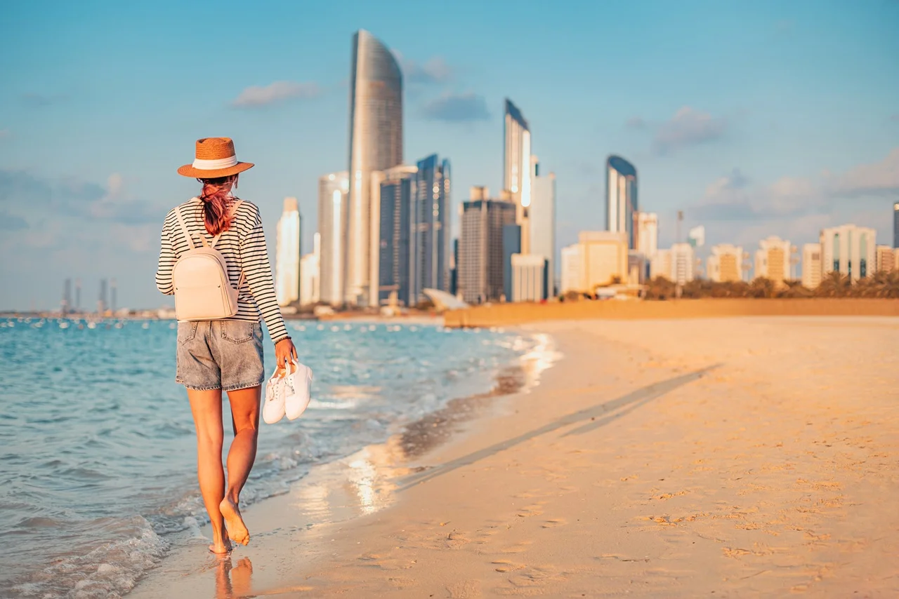 Exclusive Beaches in Abu Dhabi 2026 For Perfect Gulf Getaway 7 Young woman walking barefoot on the beach in Abu Dhabi_ carrying shoes and backpack_ enjoying