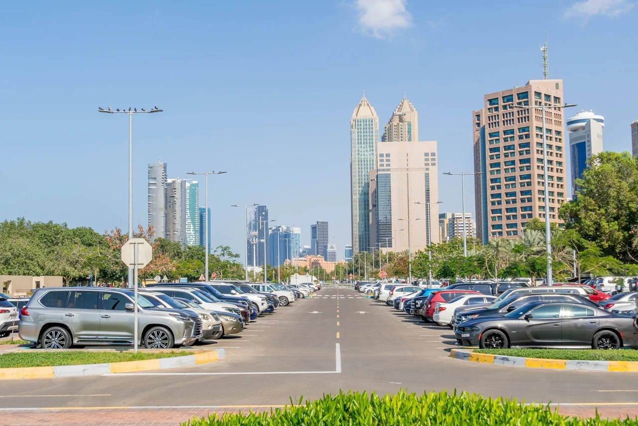 No More Taxis! 15 Trusted Car Rental Companies in Abu Dhabi 3 Wide view of a cityscape featuring tall skyscrapers under a clear blue sky. In the foreground is a parking lot lined with cars on both sides.