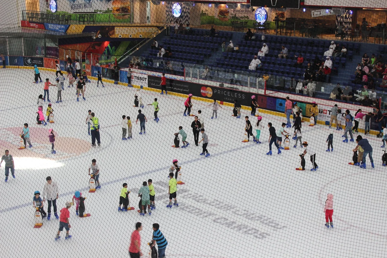 A bustling indoor ice rink with numerous skaters, including children learning to skate. Spectators watch from the stands, creating a lively atmosphere.