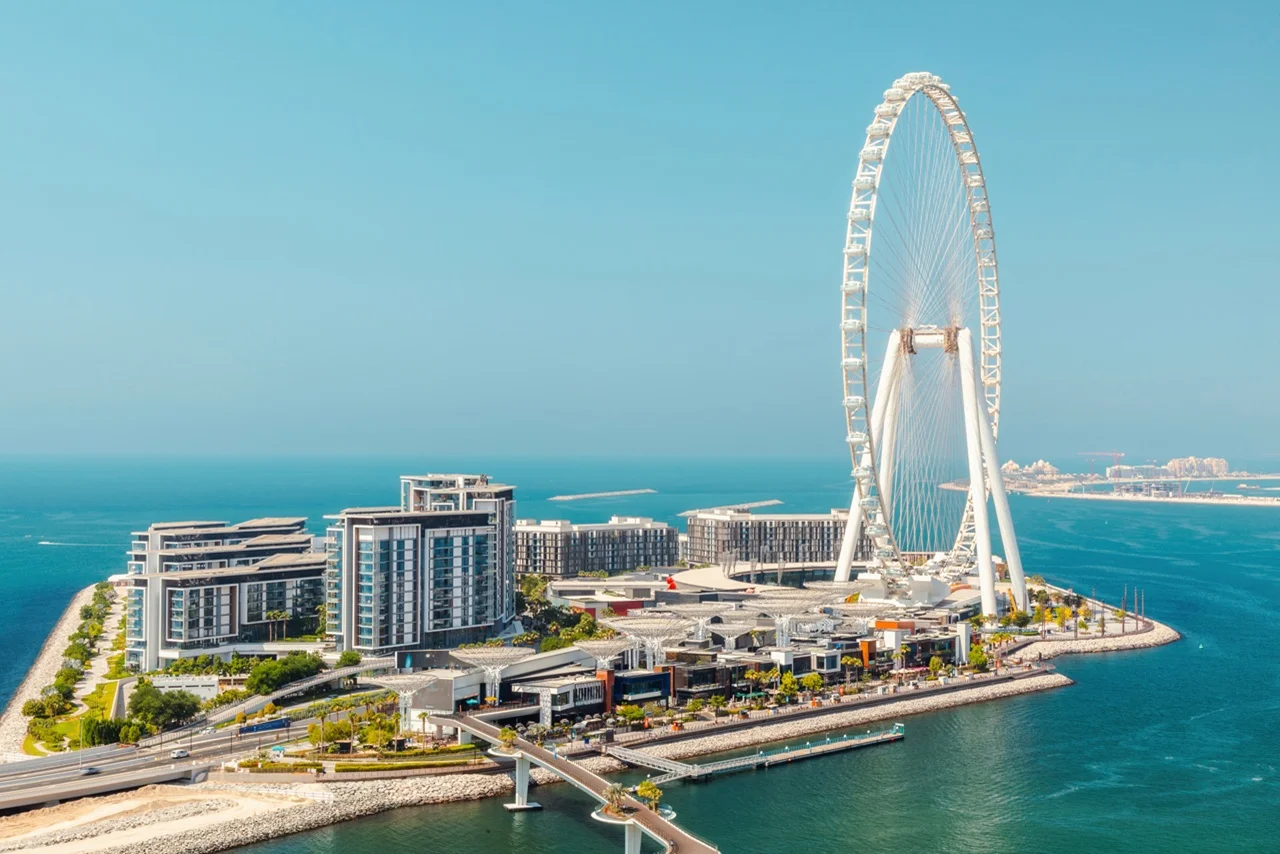 Aerial view of a coastal cityscape featuring a large Ferris wheel near modern buildings. The ocean and skyline blend under a clear blue sky, evoking tranquility.