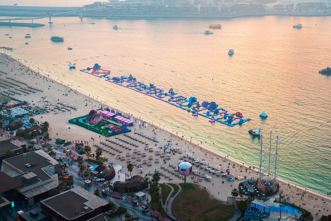 Aerial view of a crowded beach with sun loungers and umbrellas. An inflatable water park floats on calm, sunset-tinged waters, with boats nearby.