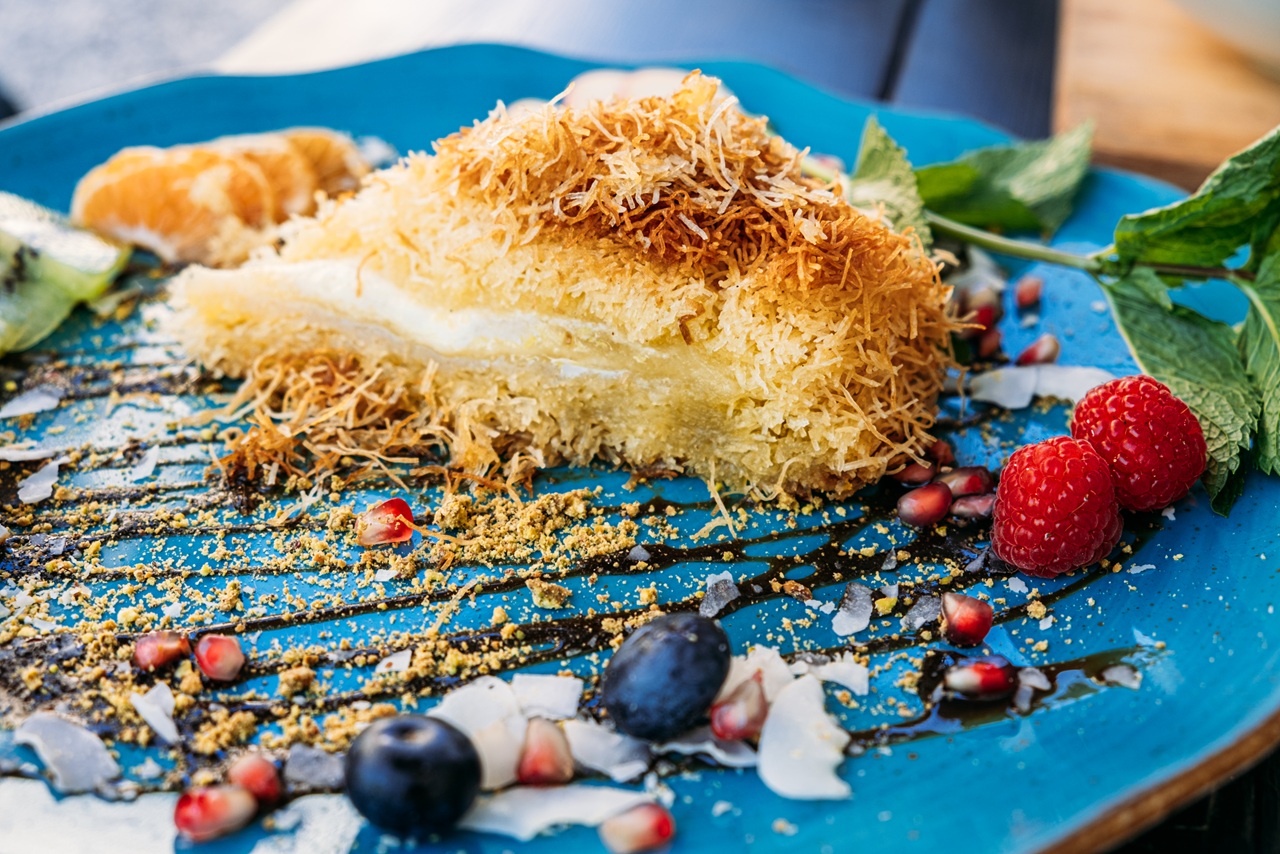A slice of cake with crispy, golden shredded pastry on a vibrant blue plate, garnished with raspberries, blueberries, mint leaves, and pomegranate seeds, creating a colorful and appetizing presentation.