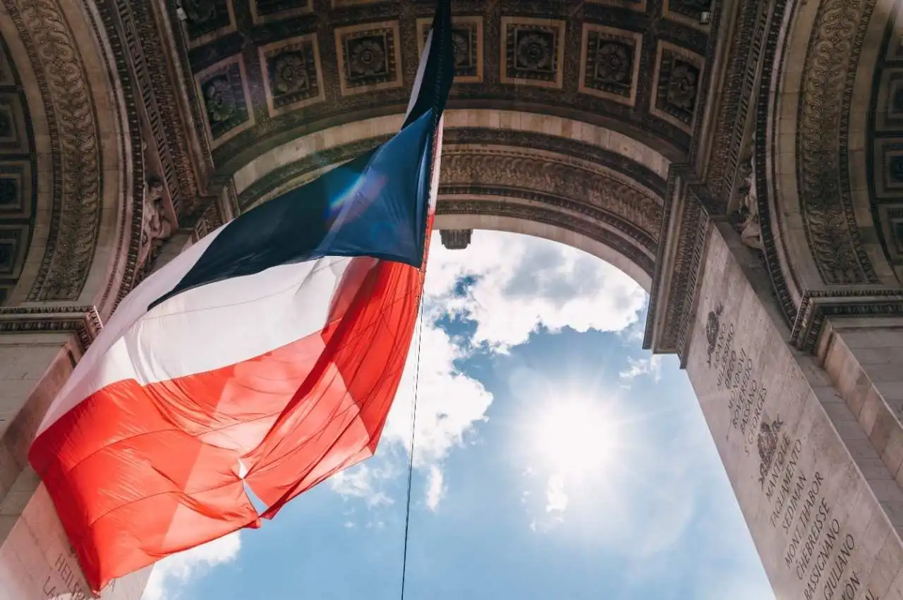 French flag waving beneath the Arc de Triomphe with sunlight shining through the arch against a blue sky.