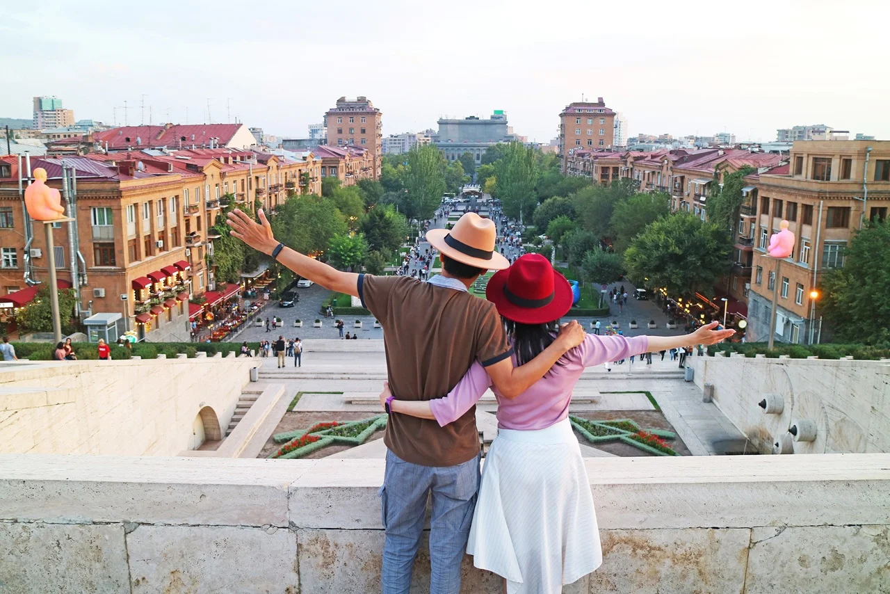 Armenia Tourist Visa for UAE Residents: Complete 2026 Travel Guide 11 A couple stands with arms outstretched, overlooking a cityscape with historic buildings, a tree-lined street, and a soft evening sky, conveying joy and exploration.