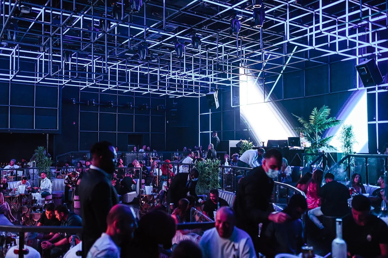A lively nightclub with dim blue and purple lighting, a geometric ceiling design, and crowds of people socializing at tables. The scene conveys an energetic, vibrant atmosphere.