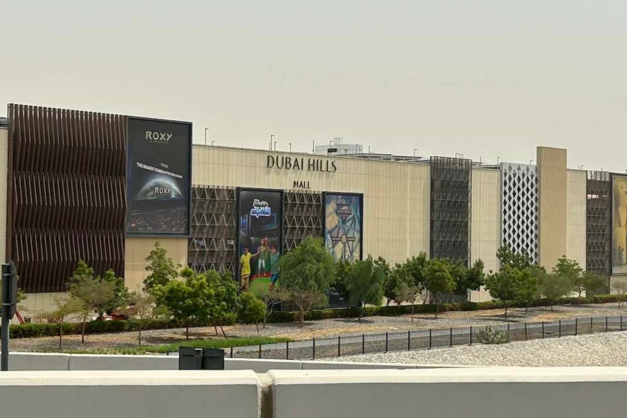 20 Best Things to Do in Dubai Hills Mall 3 Exterior of Dubai Hills Mall, featuring modern architecture with geometric patterns and large advertisements. Surrounded by greenery under a cloudy sky.