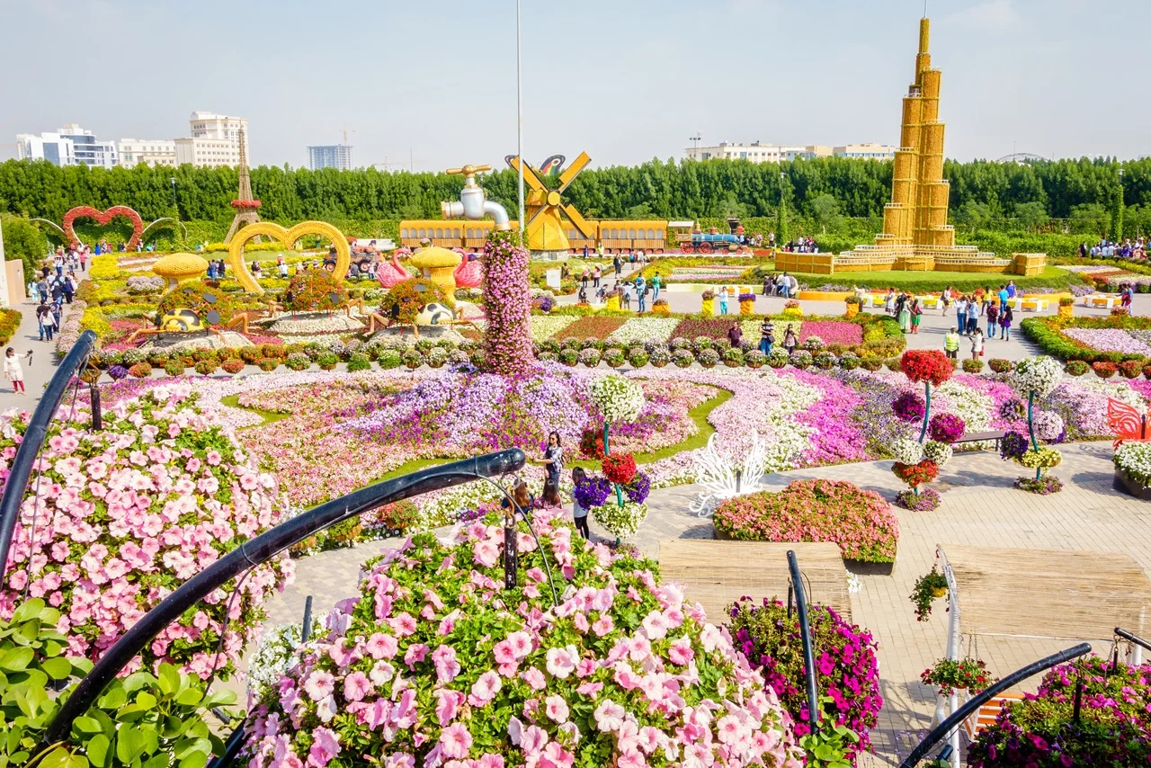 Colorful flower garden with intricate arrangements and sculptures, including a windmill and tower, under a bright blue sky. Visitors roam joyfully.
