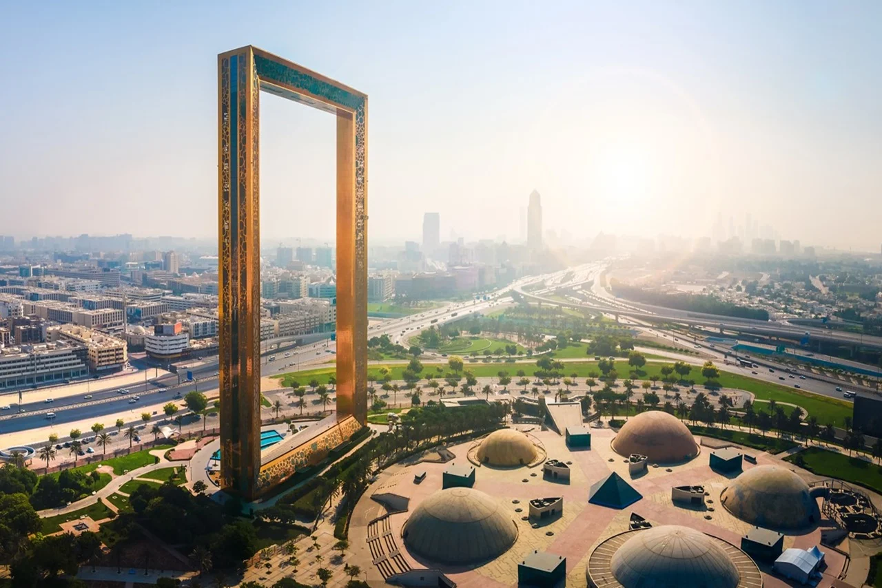A large, golden rectangular frame stands prominently against a city skyline under a bright, hazy sky. Green parks and roads surround the structure.