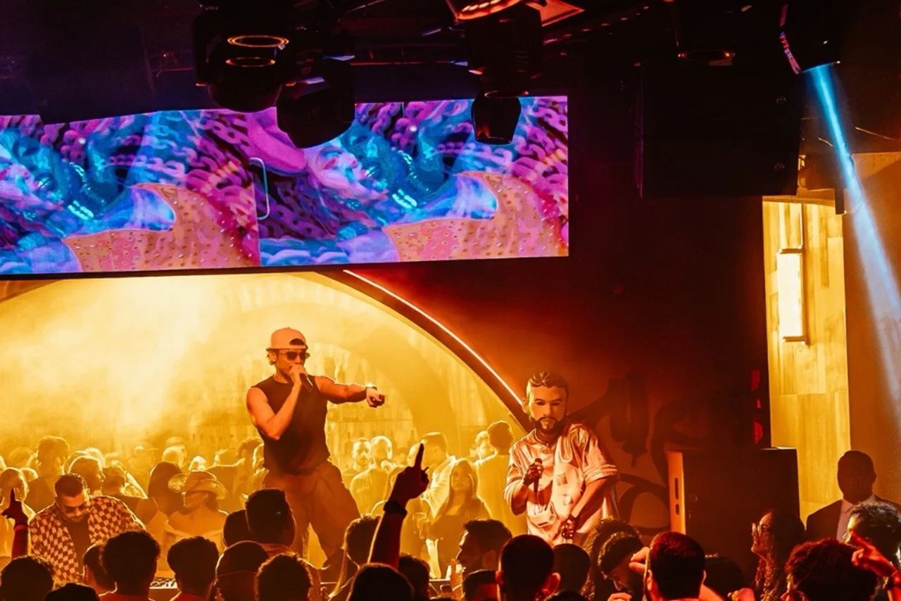 A lively nightclub scene with a performer in sunglasses and a cap engaged with a cheering crowd. Vibrant, colorful lights and digital screens create an energetic atmosphere.