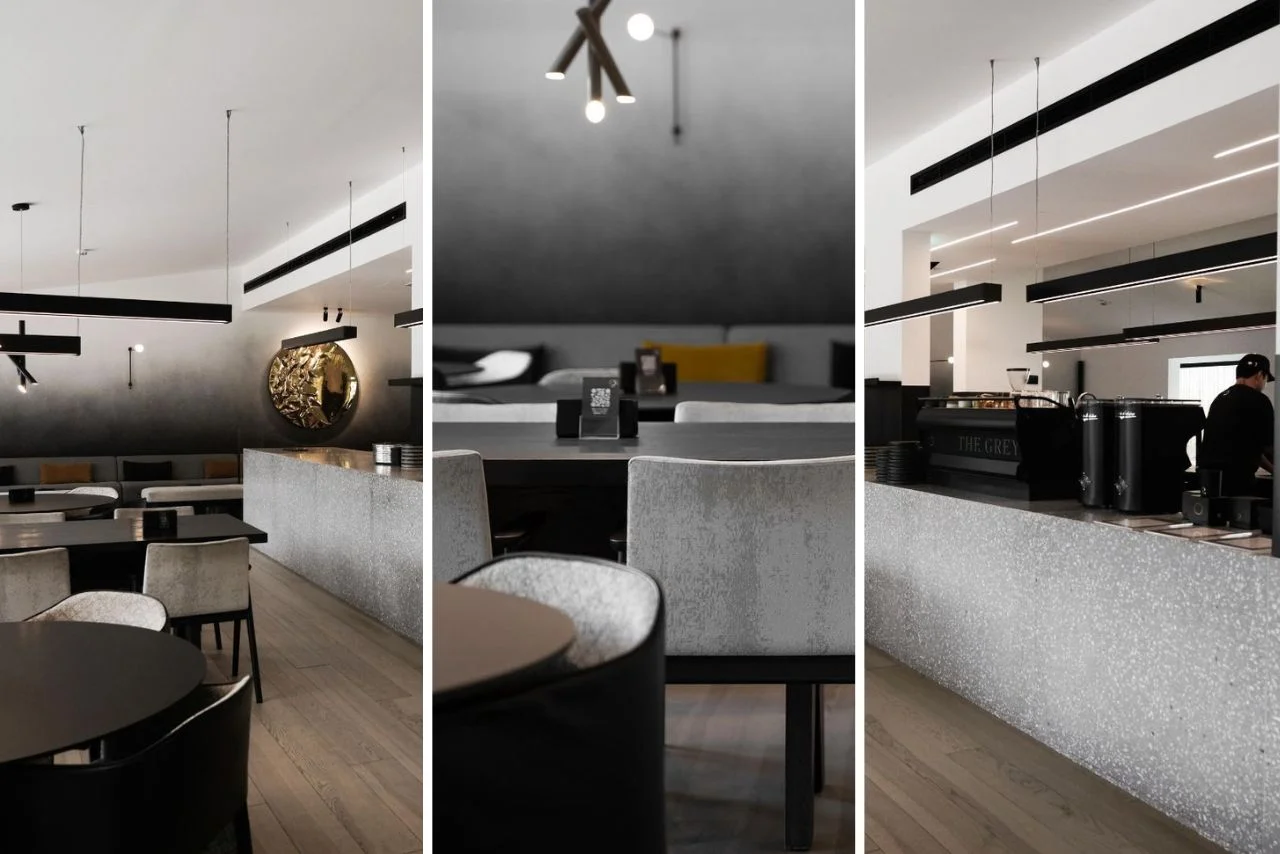 Modern cafe interior with minimalist decor. Features black tables, white chairs, hanging lights, textured walls, and a barista at a sleek bar. Cozy ambiance.