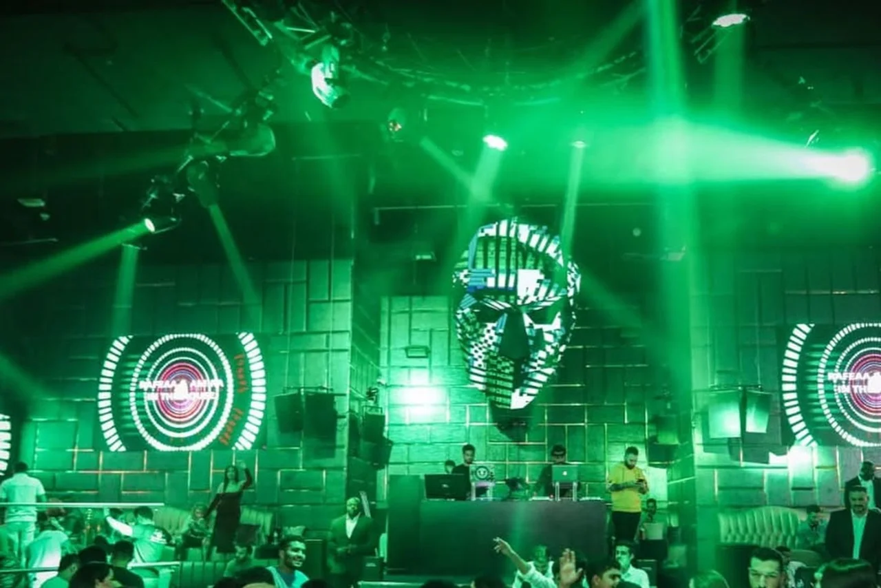 A vibrant nightclub scene with green lighting and a DJ booth featuring two large circular screens. A geometric face design decorates the wall above the crowd.