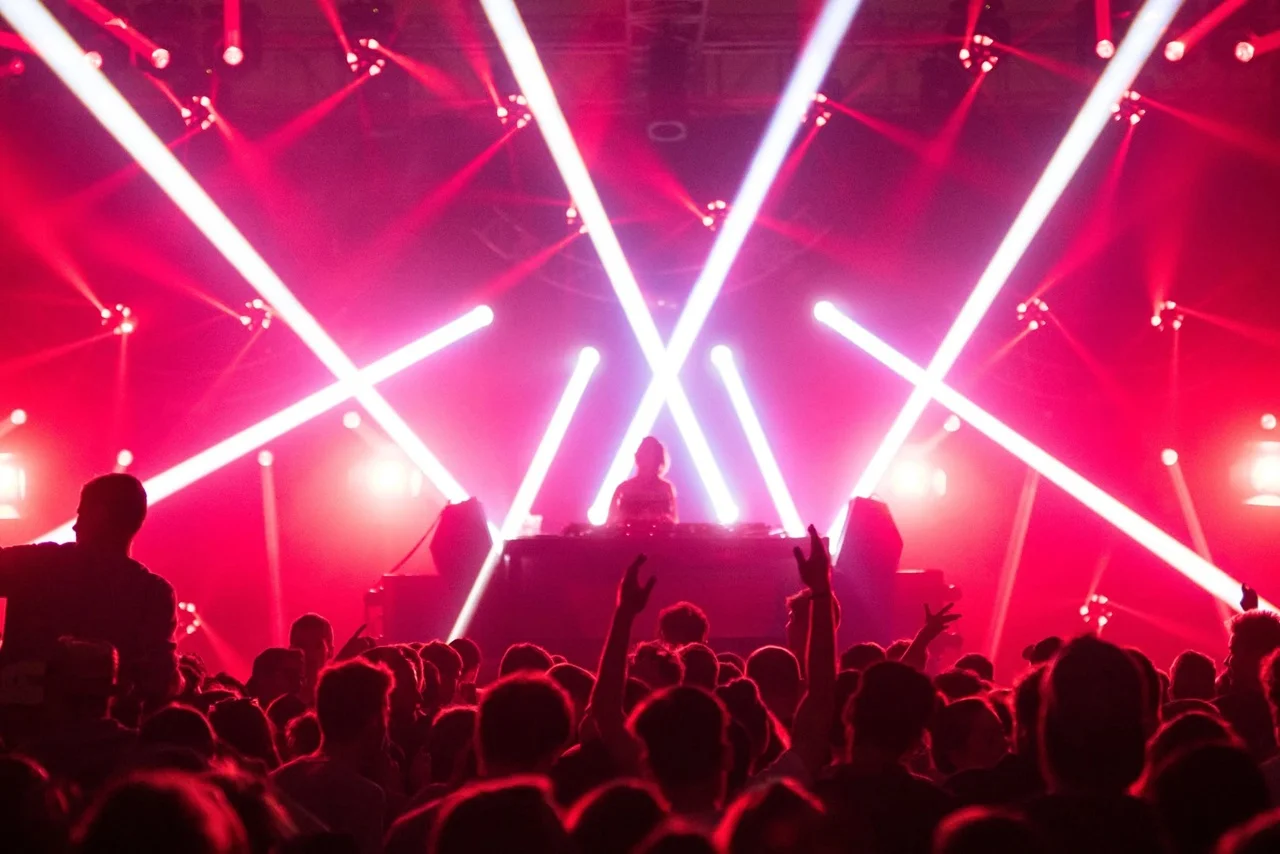 A DJ performs on stage, surrounded by vibrant red and white lights, as a crowd dances energetically, creating an electric and lively atmosphere.