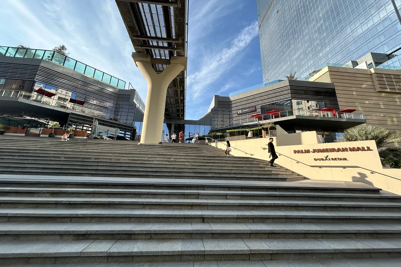 Palm Jumeirah Mall: Grand staircase at Dubai’s Polis Junction Mall, leading to modern architectural buildings with glass facades. Blue sky and people walking, indicating a lively atmosphere.