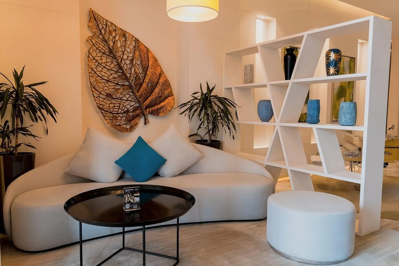 Live like a local in a fully furnished apartment or private pool suite at Arjaan by Rotana Dubai Media City 6 Modern living room with a curved white sofa, turquoise and white cushions, and a geometric bookcase. Leaf wall art and plants add elegance.