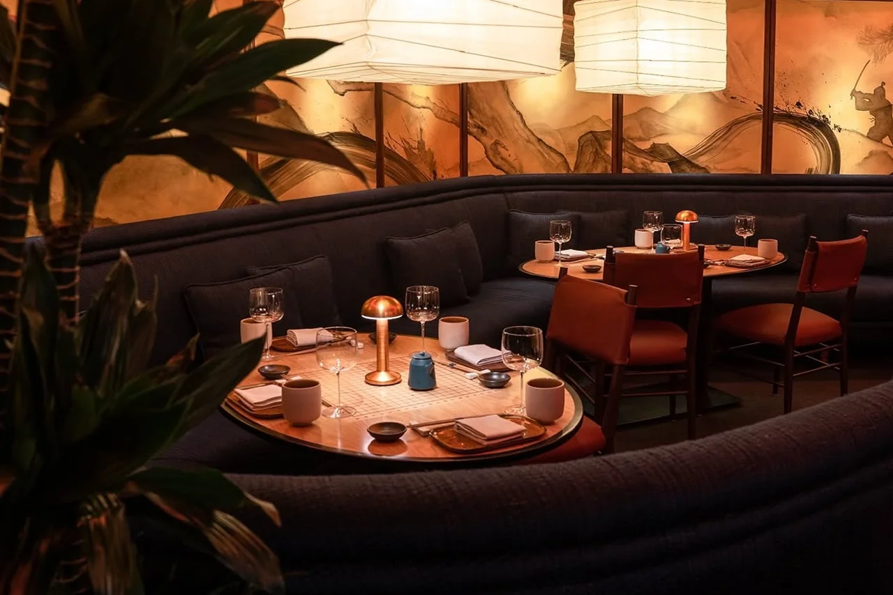 Cozy restaurant interior with a curved blue sofa and round wooden tables set for dinner. Soft lighting from large hanging lanterns creates a warm ambiance.