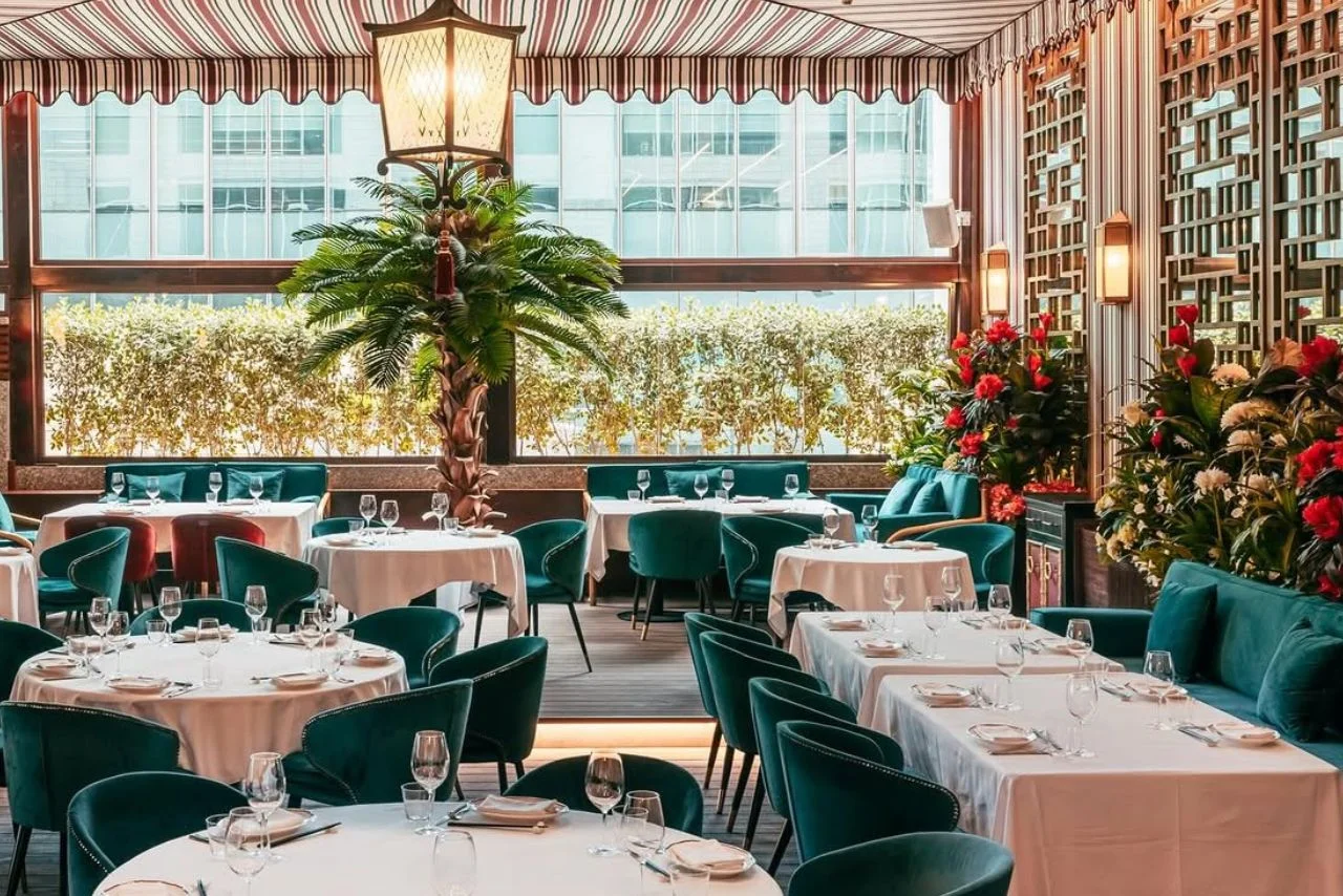 The sophisticated dining room at Shanghai Me Dubai featuring luxury teal velvet seating, neatly set dinner tables, ornate wall panels with red flowers, and large windows looking out toward city buildings.